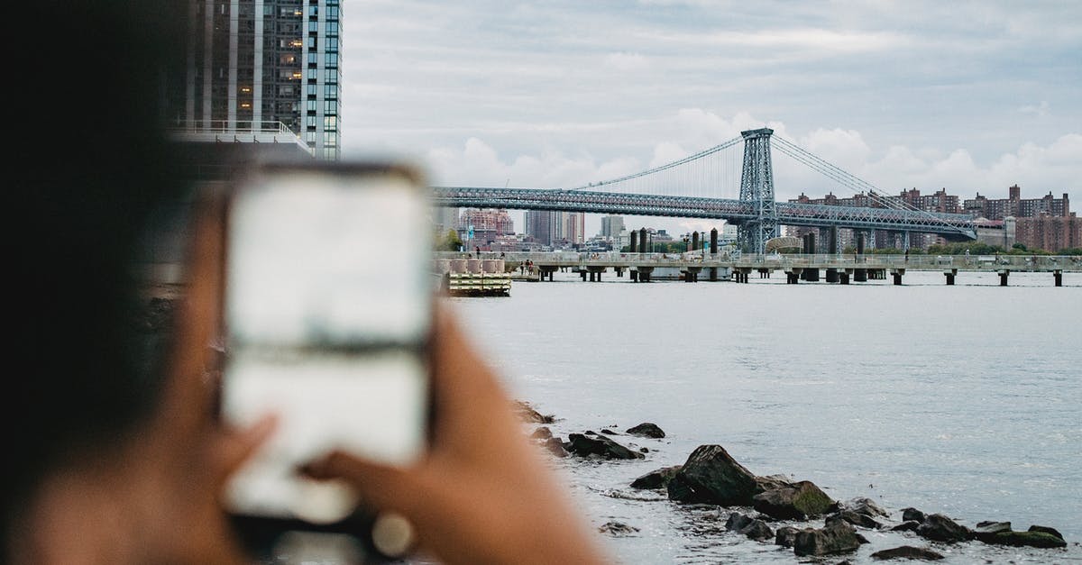 What coast or river-adjacent city is in this photo? - Crop anonymous African American male standing on embankment and taking picture of skyscrapers and bridge