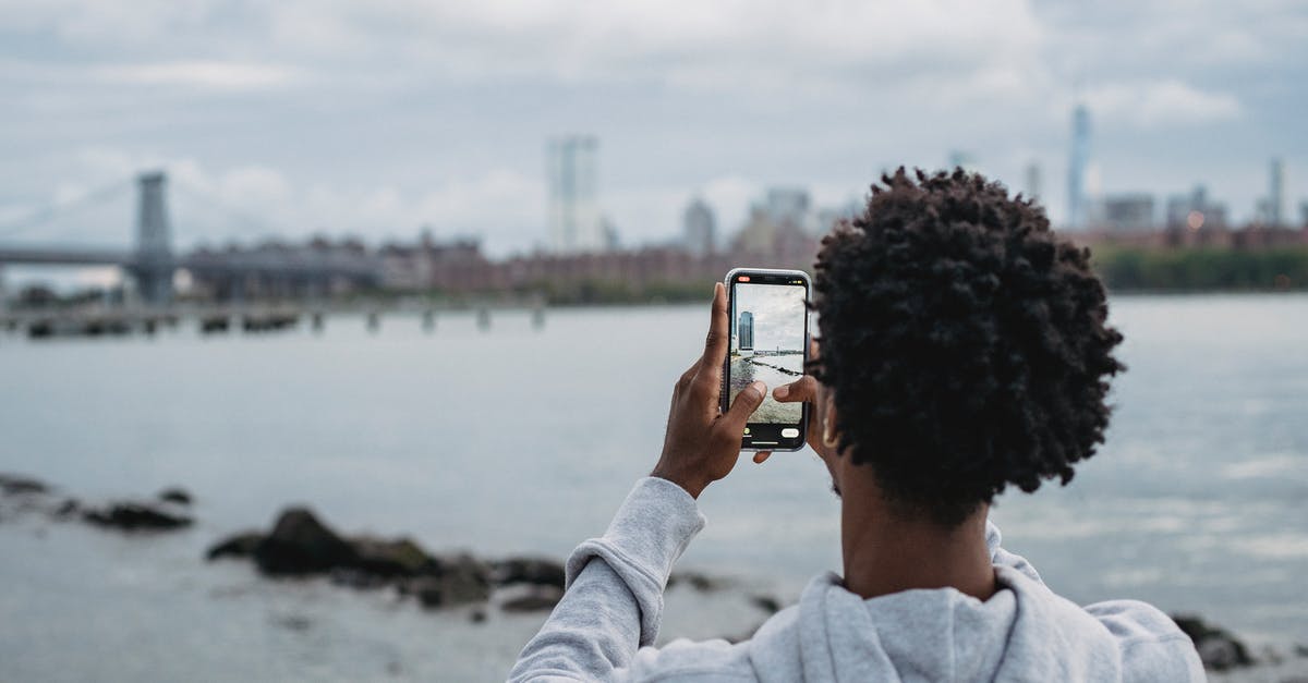 What coast or river-adjacent city is in this photo? - Black man taking photo of bridge on smartphone