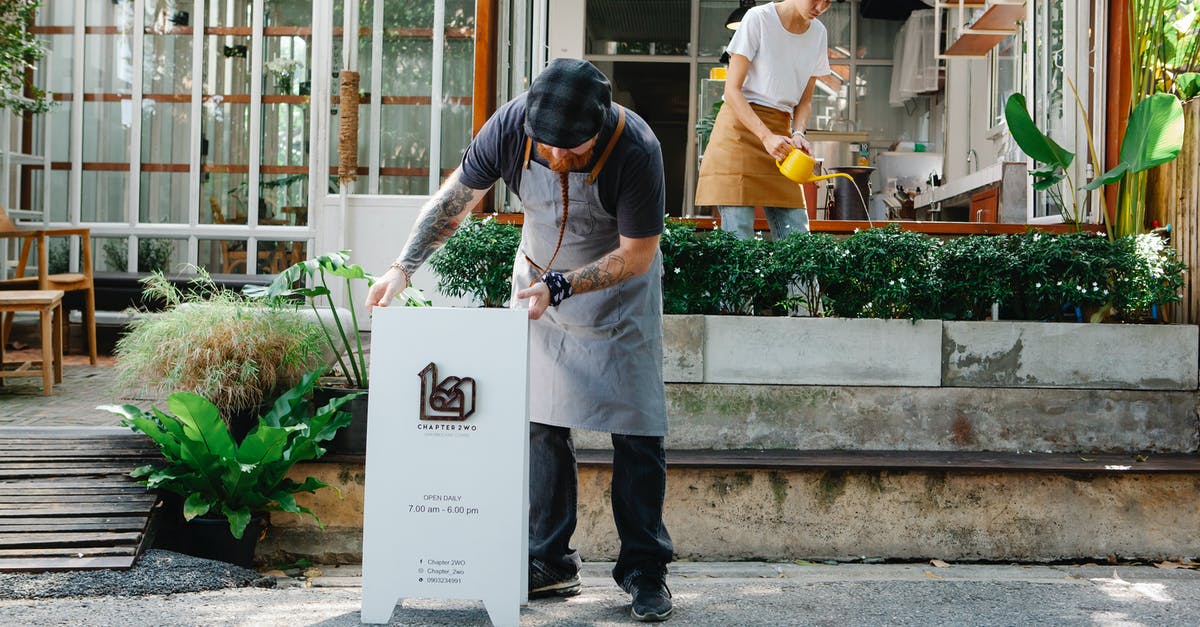 What do I put here? - Couple of coworkers in aprons and casual clothes watering plants and putting signboard near cafeteria in daytime