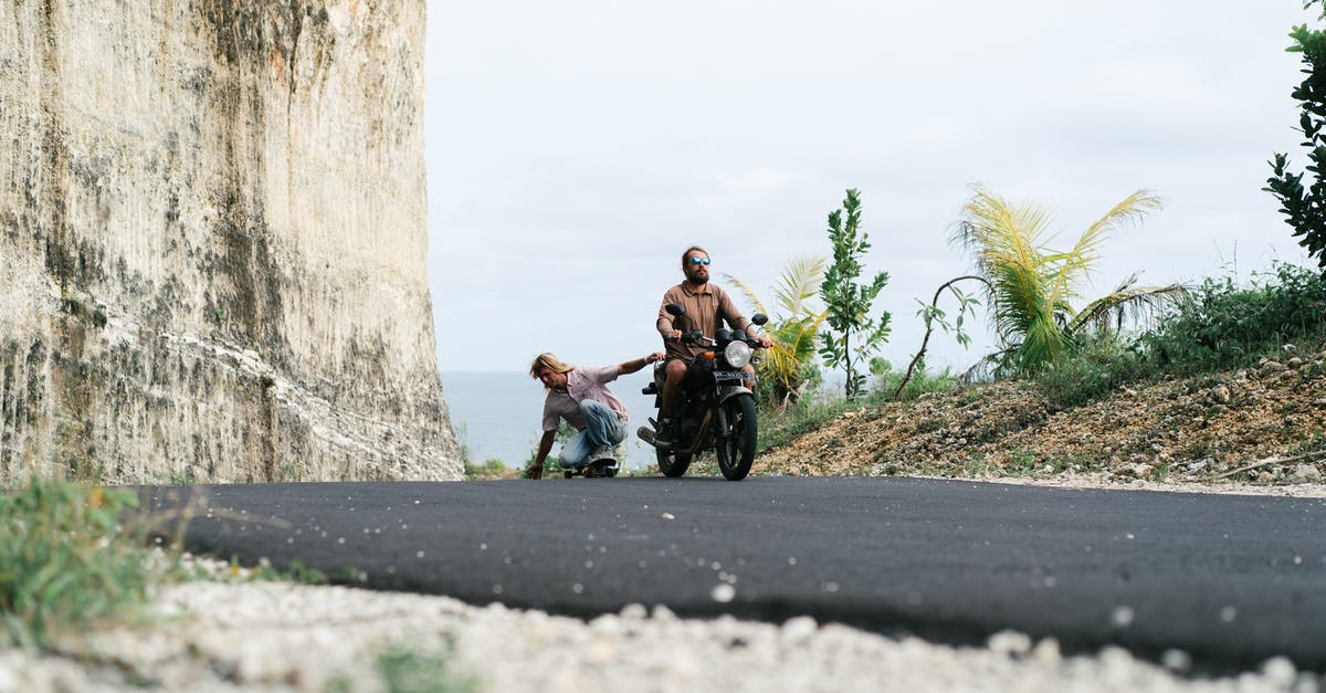 What is the experience of driving in Thailand? [closed] - Full length male driver of motorbike riding friend in sitting position on skateboard while friend holding seat and touching surface of road with another hand