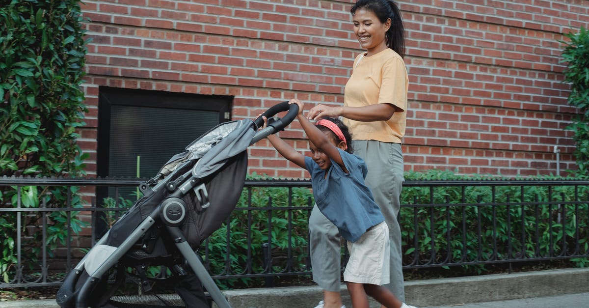 What is the name of this small town in Italy somewhere outside of Rome? - Side view of cheerful Asian woman in casual clothes walking small daughter rolling baby carriage on street near brick building in daytime