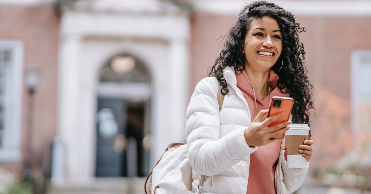 What is the rationale of SMS verification in free wifi hotspots in airports? - Cheerful young ethnic female student with long curly hair in casual clothes and backpack smiling and looking away while using mobile phone standing in campus with takeaway coffee