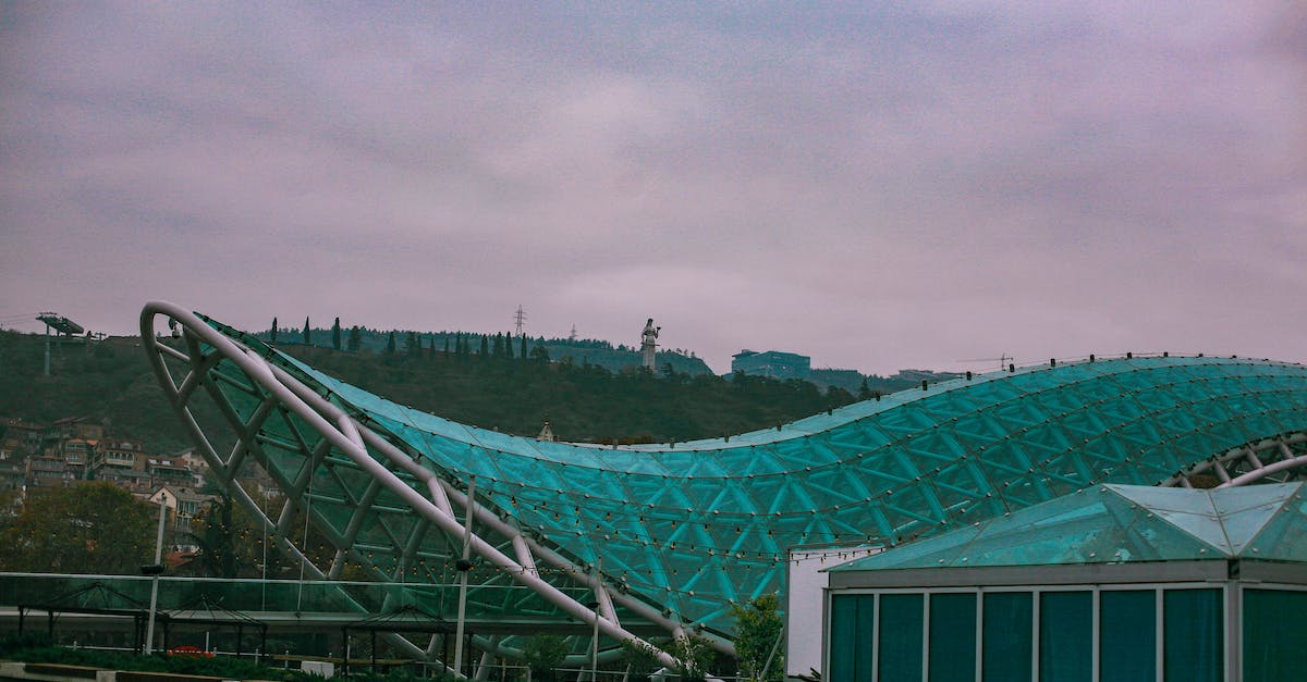 What was this ruined building in Tbilisi, Georgia that is Greek property? - Modern glass construction called Bridge of Peace and located in Tbilisi in Georgia near building and green trees and plants with hill on background under cloudy gray sky in street