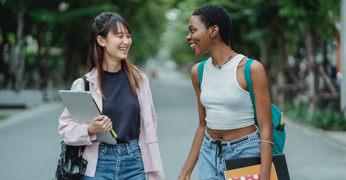 When is the best day and time to buy tickets at Qantas? - Smiling young multiethnic women standing in middle of street with laptop and notebooks while speaking with each other in summer day and wearing casual clothes while looking at each other