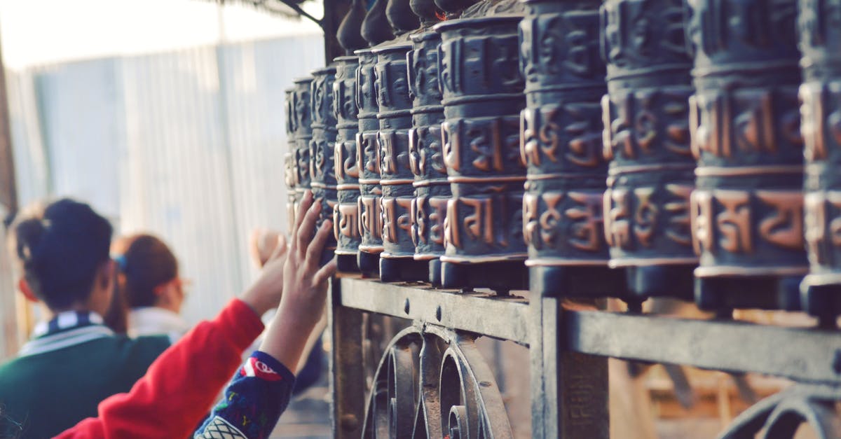 Where are the Himalayan Buddhist stupas? - Back view of anonymous tourists touching old metal Prayer wheels with hieroglyphs above ornamental fence near old Buddhist temple in Swayambhunath
