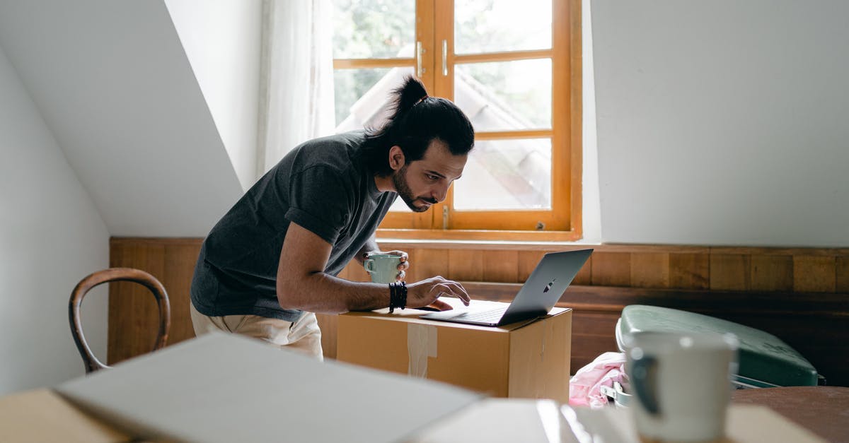 Where in Seoul can I rent a laptop? - Side view of focused bearded male in casual outfit with cup of hot drink browsing laptop while packing stuff into carton boxes