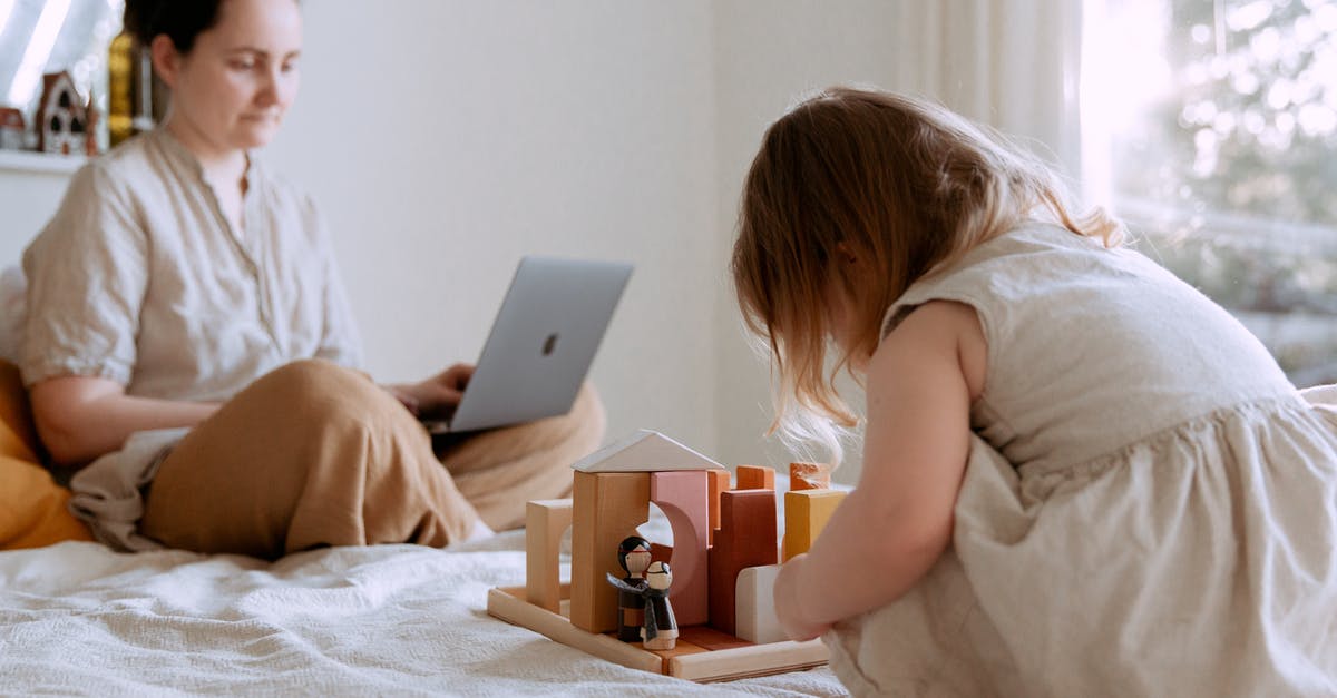 Which airlines ban the use of Knee Defenders during flight? - Side view of adorable toddler girl sitting on bed and playing with wooden toys while mother working from home with laptop on knees on sunny morning