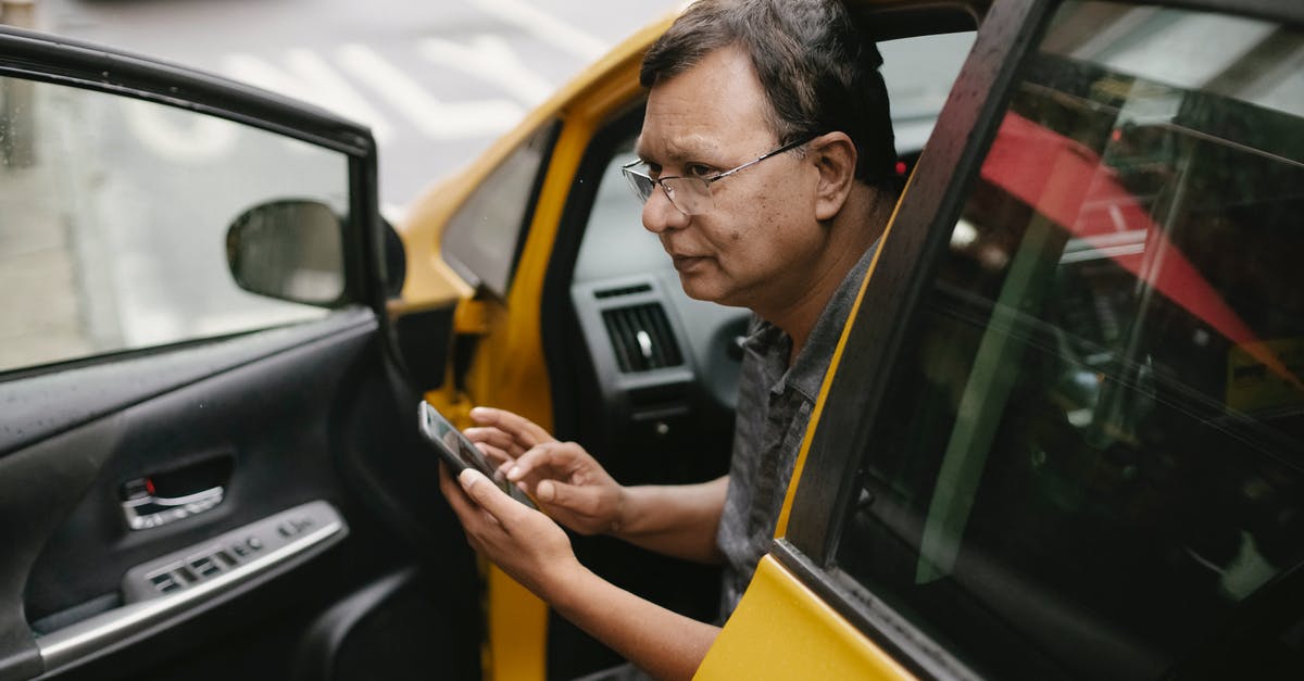 Which drivers license to use in the EU? - Crop Asian man sitting in parked yellow taxi car with open door and using smartphone while looking away