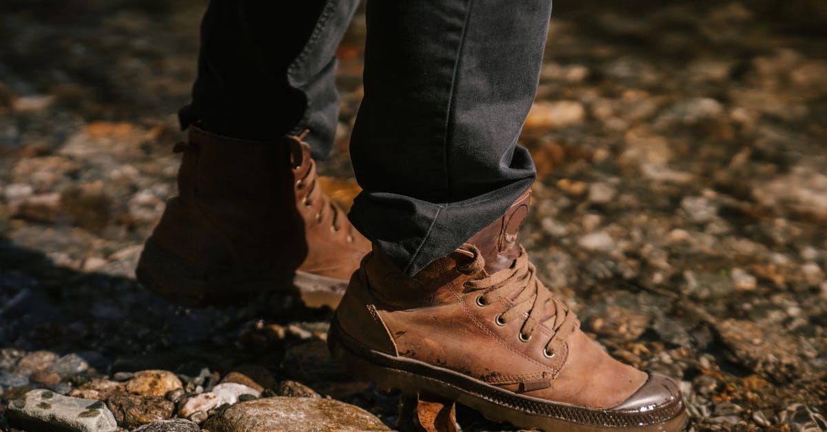 Which trekking shoes to get for trekking in Costa Rica - Crop anonymous person in brown boots and black jeans walking on stones near river bank