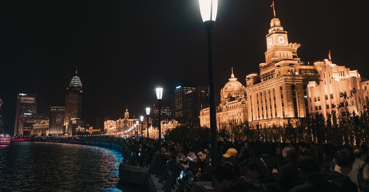 Who was this persistent person at Shanghai airport? - Low angle of aged masonry Bund exterior with clock and national flag near river and glowing lamp post above unrecognizable people in town at dusk