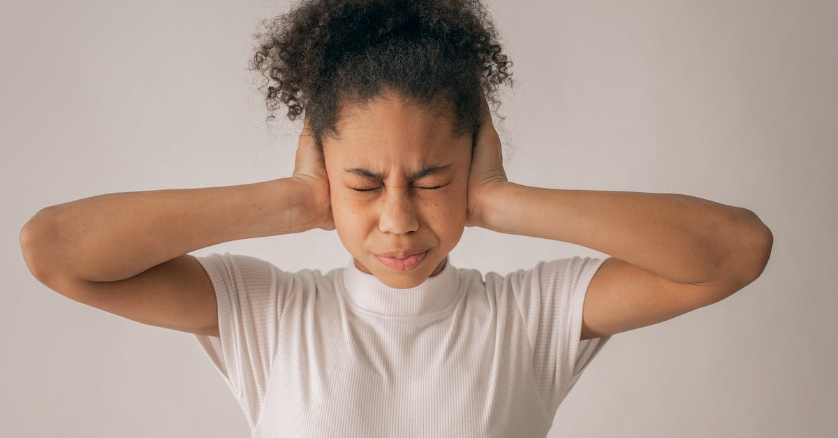 Why do I feel pressure in the ears in a plane? - Despaired African American teenage girl with black hair and closed eyes covering ears while standing on white background in light studio