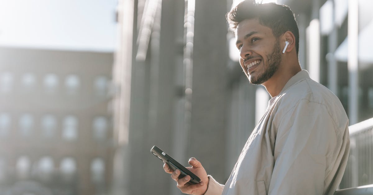 Why only earbud style earphones? - Side view of cheerful young bearded Hispanic male in TWS earbuds using mobile phone while standing near modern urban building in sunny day