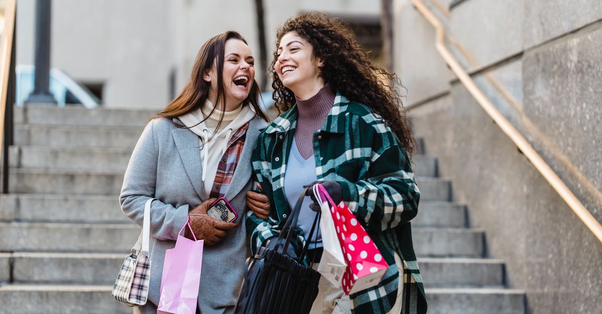 Will I have to recheck my bags? - Joyful young stylish multiracial female best friends in trendy warm clothes laughing while walking downstairs with shopping bands in hands on city street