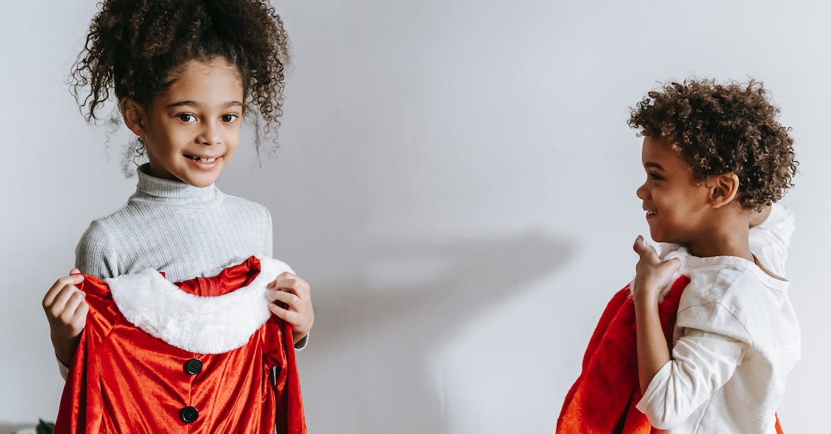 Winter apparel for Europe (November-December) - Cheerful African American siblings with red Santa Claus outfits in hands standing in light room at home during Christmas holiday