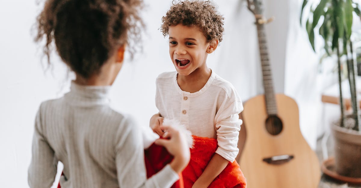 Winter apparel for Europe (November-December) - Funny African American boy looking at anonymous sister while standing with Santa Claus costumes in room with guitar during Christmas holiday